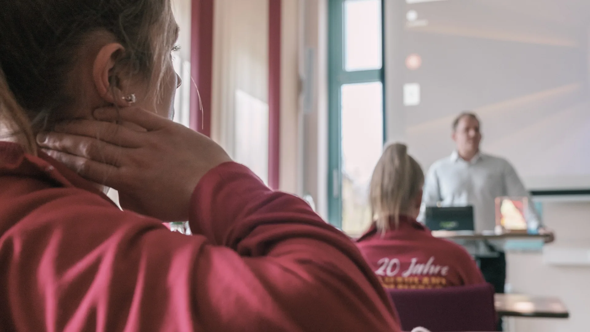 Person im dunkelroten Poloshirt von hinten, die Hand im Nacken; dahinter ein heller Schulungsraum, in dem ein Referent vor der Sitzgruppe steht