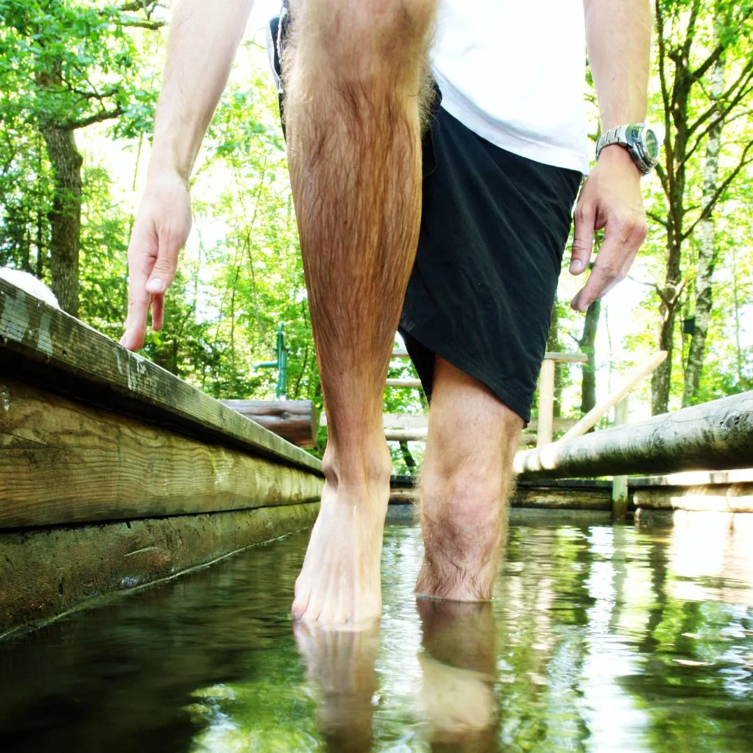 Person geht barfuß durch ein Wassertretbecken im Freien und stützt sich an einem Holzgeländer ab