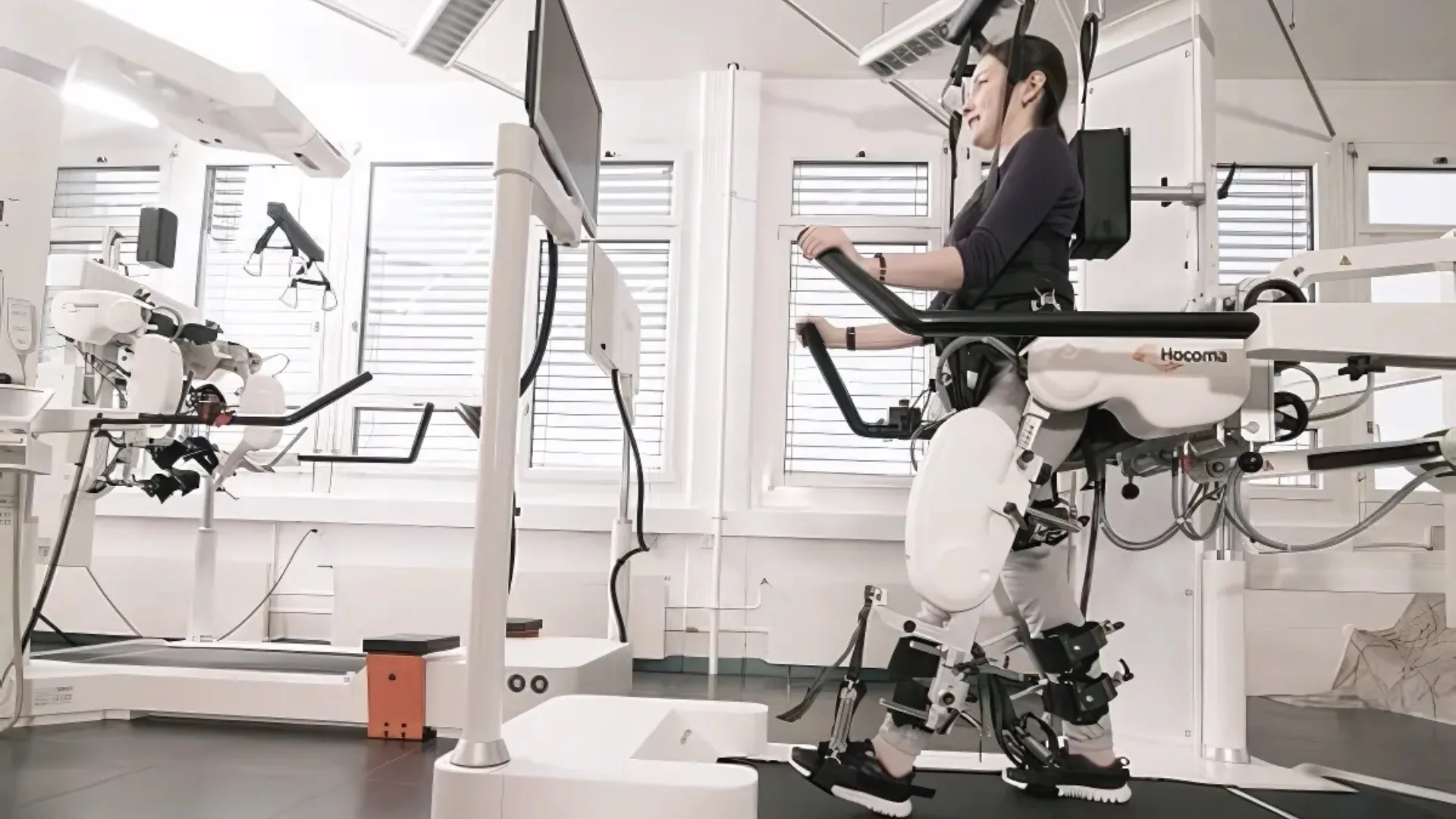 Therapieraum mit robotikgestützten Geräten, im Hintergrund steht eine Patientin in einem Stehtrainer