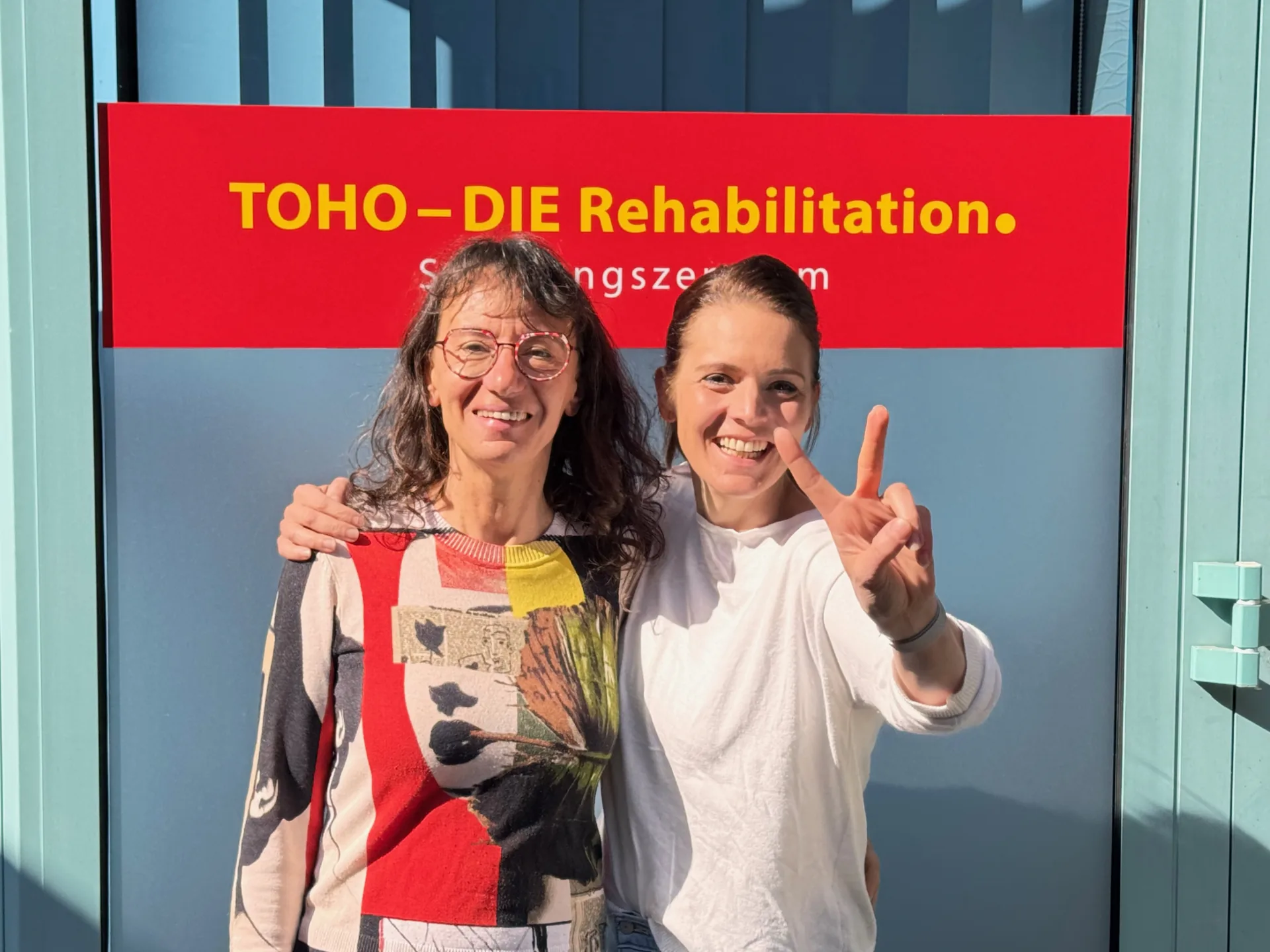 Zwei Kursteilnehmerinnen stehen lachend vor dem roten Banner „TOHO – DIE Rehabilitation. Schulungszentrum“
