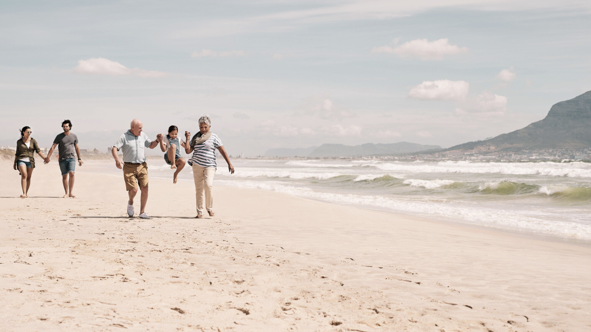 Drei Generationen einer Familie gehen Hand in Hand am Strand, ein Kind springt in der Mitte hoch