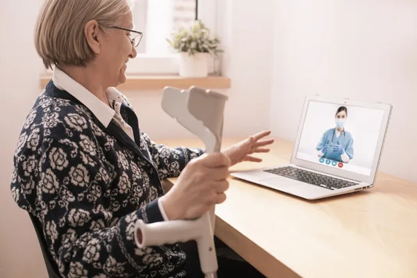 Ältere Patientin im Videogespräch mit einer Therapeutin, in den Händen eine Unterarmgehstütze